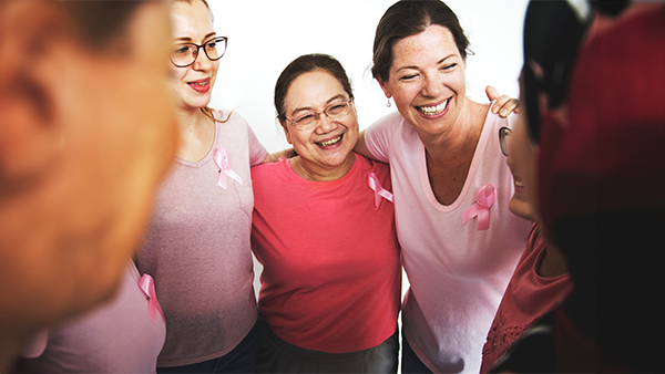 A group of five women in pink clothing, with pink ribbon pins, standing together and smiling.